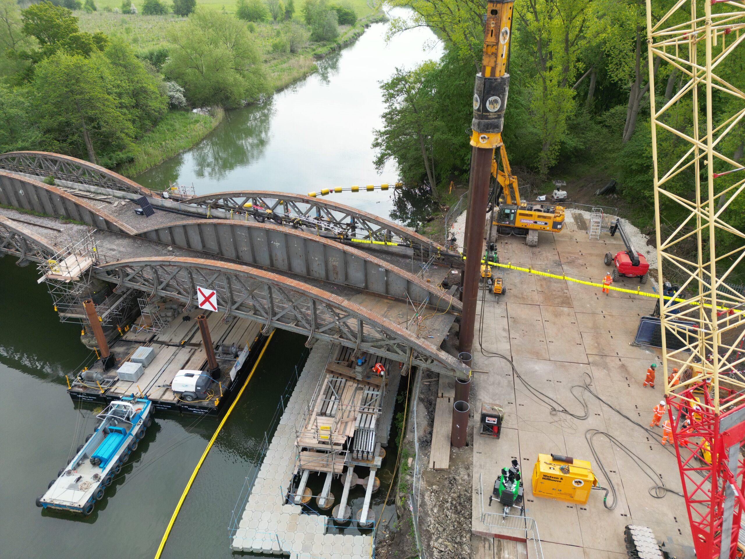 Image shows a bridge spanning the river in Abingdon-on-Thames. Also pictured are a Watson & Hillhouse BSP CX hydraulic hammer with acoustic shrouding and power pack. The hammer is installing 20m tube piles as supports for the new bridge.